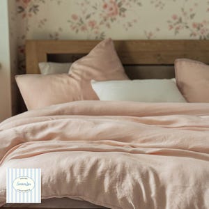 May include: A bed dressed with a light pink linen duvet cover and matching pillowcases. A white pillow is centered on the bed. The headboard is wooden, and the wallpaper has a floral pattern. The logo on the bottom left says "Sensory Luxe".