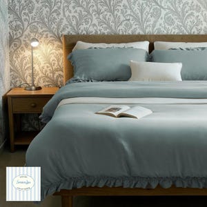 May include: A bed dressed with a teal duvet cover and pillows. A white pillow is centered on the bed. A small wooden bedside table with a lamp is to the left. An open book rests on the duvet. The wallpaper has a floral design.