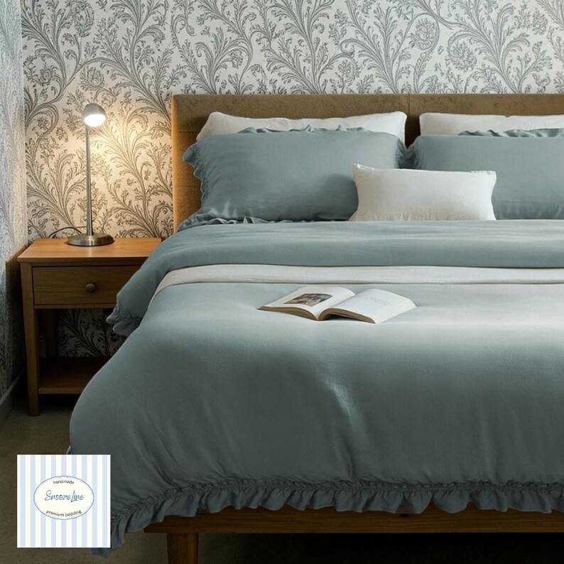 May include: A bed dressed with a teal duvet cover and pillows. A white pillow is centered on the bed. A small wooden bedside table with a lamp is to the left. An open book rests on the duvet. The wallpaper has a floral design.