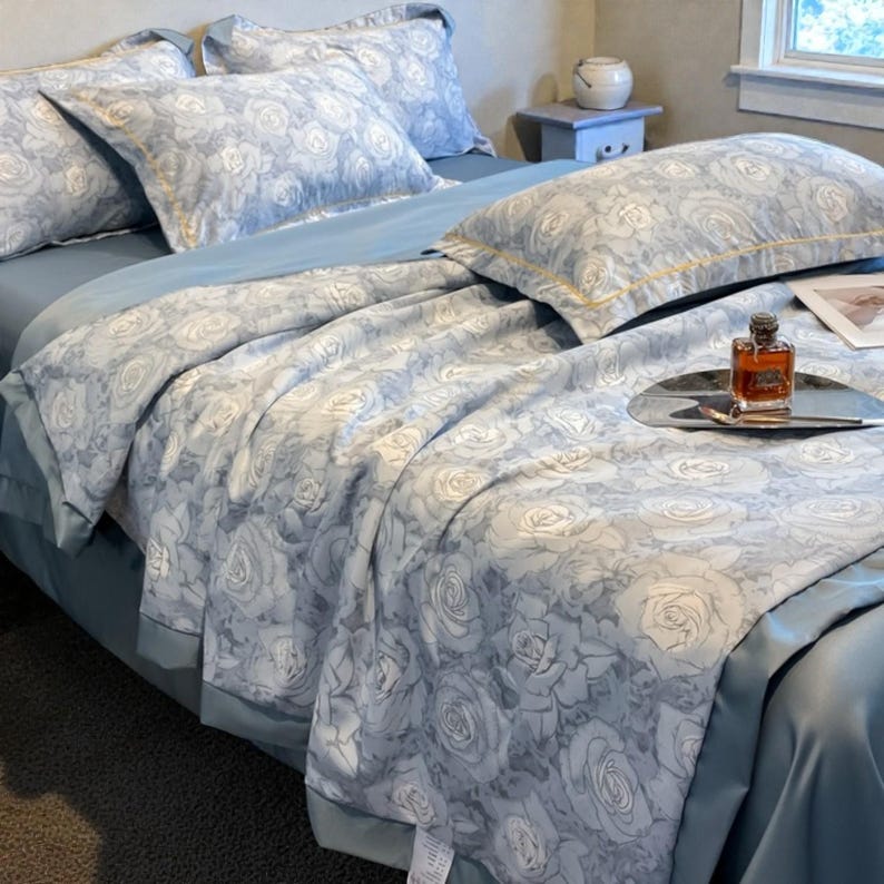 May include: A bed with light blue sheets and a comforter featuring a white rose pattern. Matching pillowcases are visible, along with a decorative tray holding a perfume bottle. The bedding has a luxurious appearance.