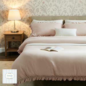 May include: A bed dressed in light pink bedding, including a comforter and pillows. A white pillow is placed centrally. A small wooden bedside table with a lamp is to the left. The wallpaper features a floral pattern. An open book lies on the comforter.