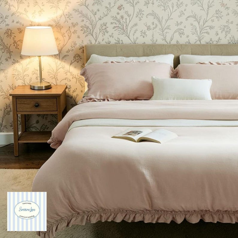May include: A bed dressed in light pink bedding, including a comforter and pillows. A white pillow is placed centrally. A small wooden bedside table with a lamp is to the left. The wallpaper features a floral pattern. An open book lies on the comforter.