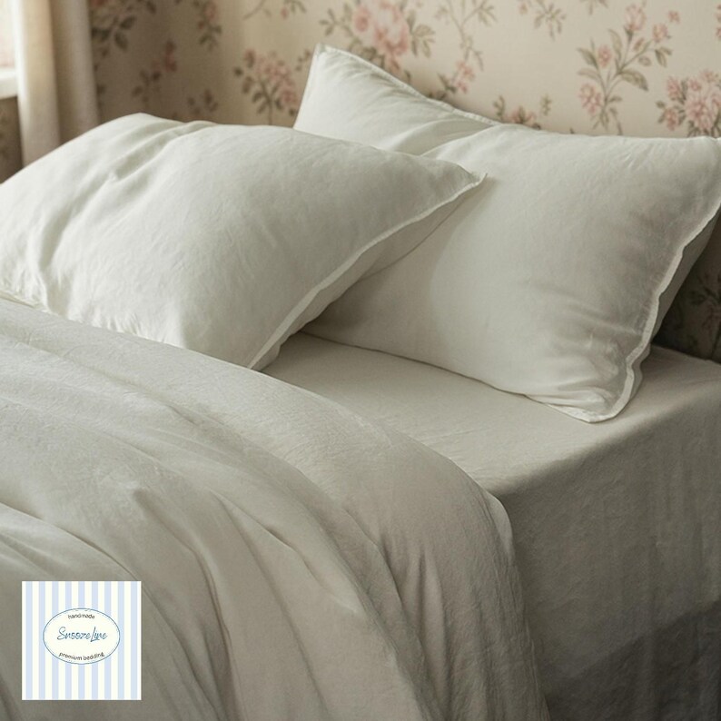 May include: Off-white bedding set with two pillows and a folded duvet. The pillows have a subtle border detail. The bedding is arranged on a bed, with a floral wallpaper background. The image includes a logo with the text "Sensore Luxe premium bedding".