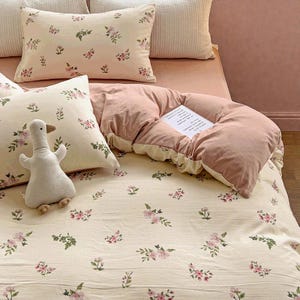 Peut inclure: Une parure de lit avec un motif floral rose et vert sur fond crème. L'ensemble comprend des oreillers, une couette et un jouet en forme de canard. La couette est de couleur rose uni.