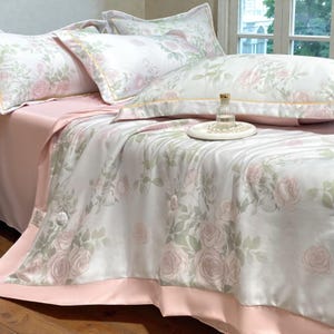 May include: A luxurious bedding set featuring a floral rose pattern in soft pink and green hues on a white background. The set includes pillows, a comforter, and a pink sheet. A small glass bottle and a tray with pearls are on the bed.