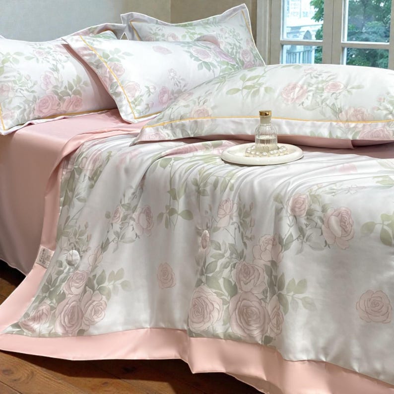 May include: A luxurious bedding set featuring a floral rose pattern in soft pink and green hues on a white background. The set includes pillows, a comforter, and a pink sheet. A small glass bottle and a tray with pearls are on the bed.