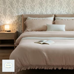 May include: A bed dressed in blush-pink bedding, including a duvet cover with a ruffled edge and matching pillows. An open book lies on the duvet. A bedside table with a lamp is visible. The wallpaper has a floral pattern.