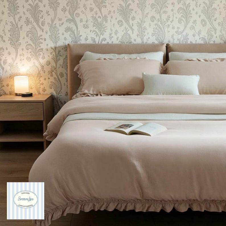May include: A bed dressed in blush-pink bedding, including a duvet cover with a ruffled edge and matching pillows. An open book lies on the duvet. A bedside table with a lamp is visible. The wallpaper has a floral pattern.