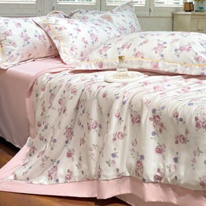 May include: A bed with a pink sheet set and a floral patterned comforter and pillowcases. The comforter and pillowcases have a white background with pink and purple roses. The pillowcases have a yellow trim.