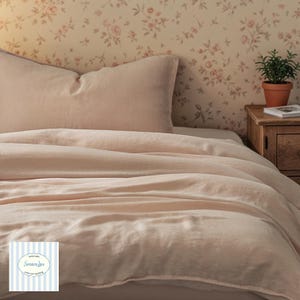 May include: A bed dressed with a blush-coloured linen duvet cover and pillow. The bedding has soft folds and a relaxed look. A small potted plant sits on a wooden bedside table. The wall behind the bed is decorated with floral wallpaper.