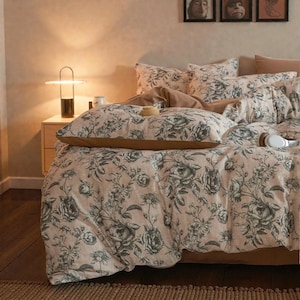 以下が含まれることがあります： 花柄の掛け布団カバーと枕カバーがセットされたベッド。淡いピンク色の背景に、グレーのバラと葉が描かれています。ベッドには茶色のベッドスカートと枕のアクセントも付いています。モダンなランプが木製のナイトスタンドの上に置かれています。