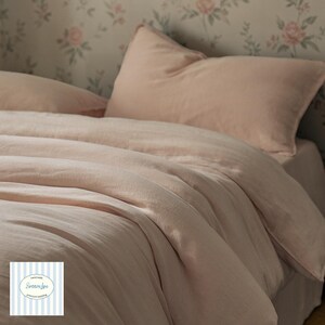 May include: A bed dressed in light pink bedding, including a duvet cover and pillowcases. The bedding has a soft, textured appearance. The background features a floral wallpaper with pink roses.