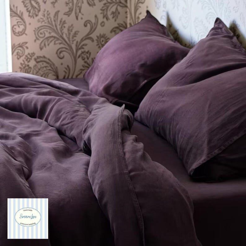 May include: A bed dressed in deep purple bedding, including a fitted sheet, duvet, and two pillows. The fabric has a soft, wrinkled texture. The background features patterned wallpaper. A logo is visible in the bottom left corner.