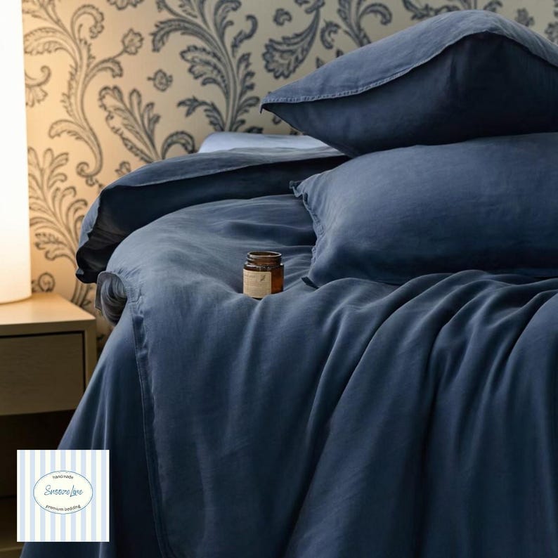 May include: A navy blue linen bedding set, including a duvet cover and pillowcases, arranged on a bed. A small brown jar sits on the bed. The background features a floral wallpaper and a bedside table.