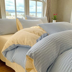 May include: A bed dressed with a light blue and yellow duvet cover set. The bedding features a ribbed texture and is complemented by matching pillows. The bed is positioned near a window offering a view of a lake and mountains.