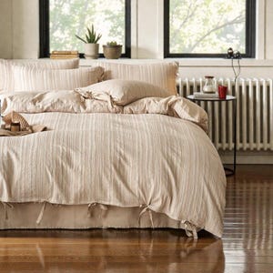 Puede incluir: Un juego de cama beige a rayas, que incluye una funda nórdica, fundas de almohada y una falda de cama. La funda nórdica y las fundas de almohada tienen un patrón de rayas verticales. La falda de cama es de color beige liso y atada con cintas.