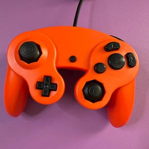 May include: An orange video game controller with black buttons and a black joystick. The controller has a black cord attached. The controller is on a purple background.