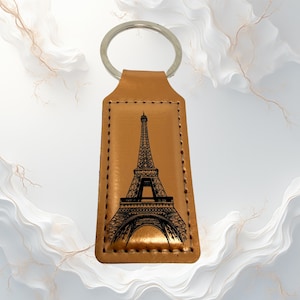 May include: Tan leather keyring with a black illustration of the Eiffel Tower. The keyring has a silver metal ring and a rectangular shape with stitched edges. The Eiffel Tower design is centered on the leather.