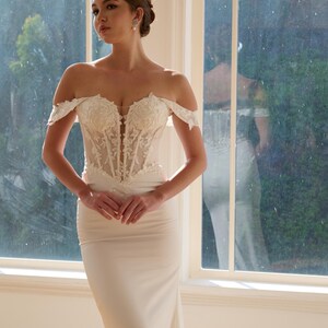 May include: Off-the-shoulder ivory wedding gown with a fitted bodice and a flowing skirt. The bodice features intricate floral lace detailing. The dress is floor-length and has a classic, elegant design, perfect for a wedding.