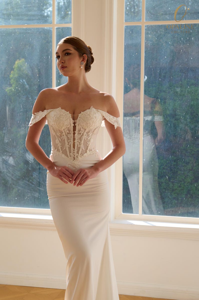 May include: Off-the-shoulder ivory wedding gown with a fitted bodice and a flowing skirt. The bodice features intricate floral lace detailing. The dress is floor-length and has a classic, elegant design, perfect for a wedding.