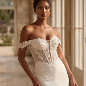 May include: A white off-the-shoulder wedding dress with a fitted bodice and a flowing skirt. The bodice features intricate lace detailing and a sheer panel. The dress is worn by a person with dark hair styled in an updo, standing in a well-lit hallway.