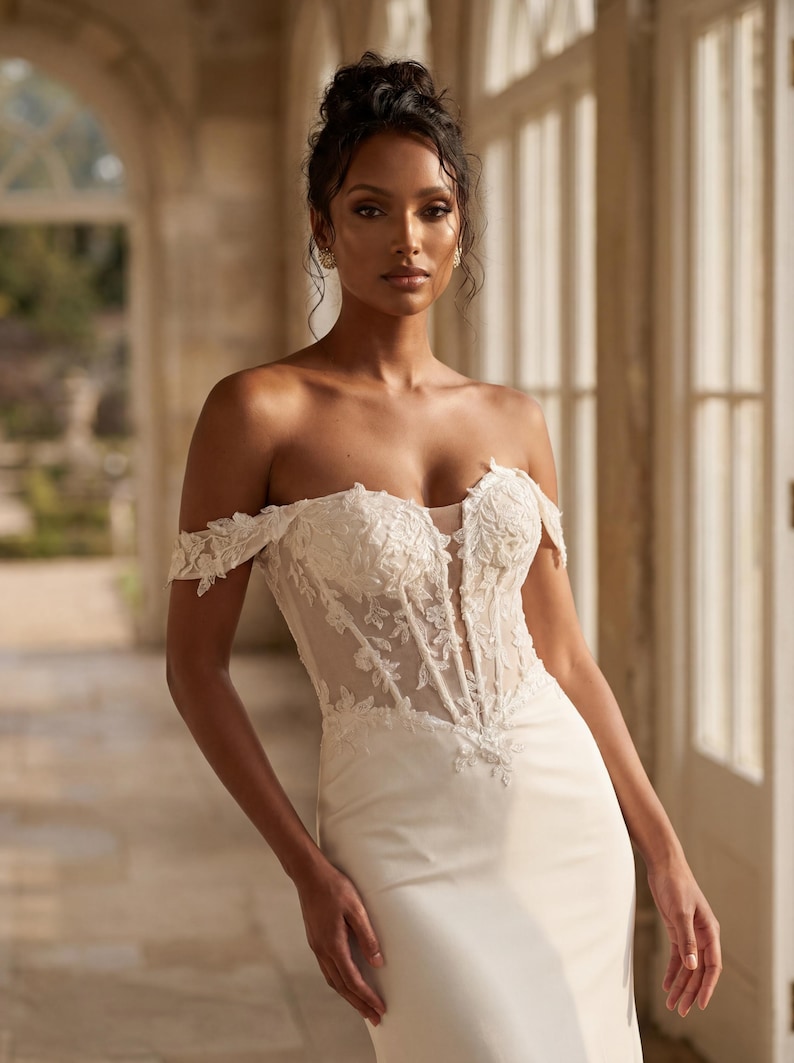May include: A white off-the-shoulder wedding dress with a fitted bodice and a flowing skirt. The bodice features intricate lace detailing and a sheer panel. The dress is worn by a person with dark hair styled in an updo, standing in a well-lit hallway.