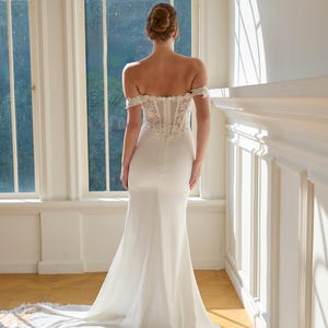 May include: A white off-the-shoulder wedding gown with a fitted bodice and a flowing skirt. The dress features lace detailing on the bodice and a long train with lace trim. The Opaline Couture logo is visible in the upper right corner.