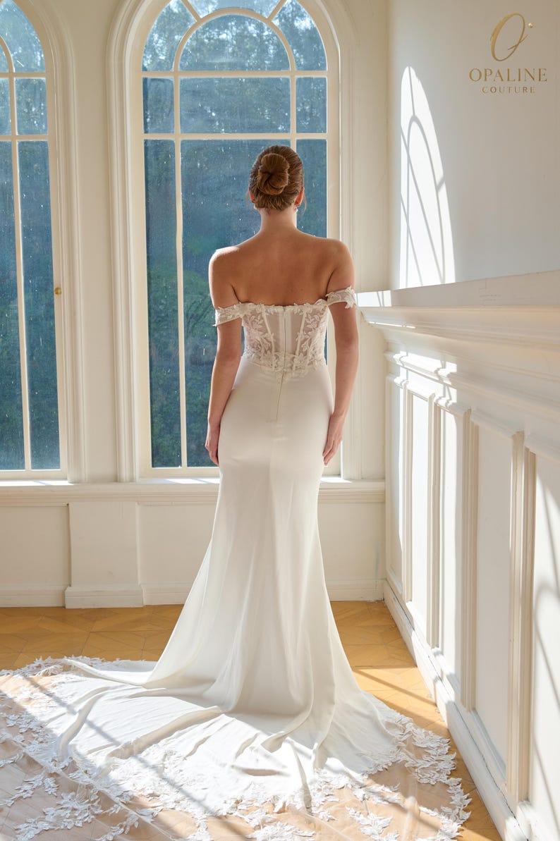 May include: A white off-the-shoulder wedding gown with a fitted bodice and a flowing skirt. The dress features lace detailing on the bodice and a long train with lace trim. The Opaline Couture logo is visible in the upper right corner.