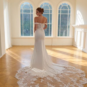 May include: A white off-the-shoulder wedding gown with a long train. The dress features a fitted bodice with lace detailing and a flowing skirt. The train is adorned with delicate floral lace. The image is set in a bright room with large arched windows.