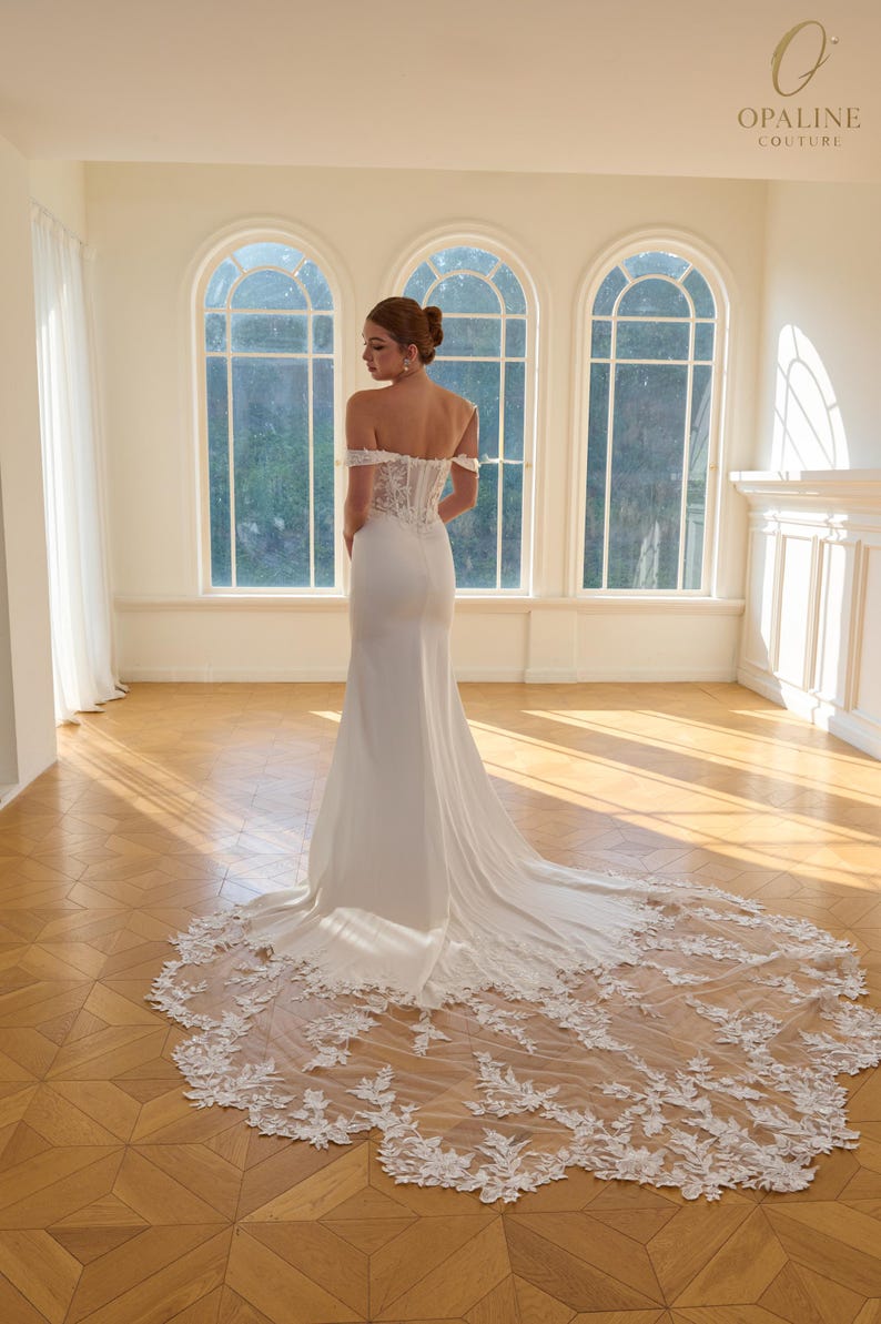 May include: A white off-the-shoulder wedding gown with a long train. The dress features a fitted bodice with lace detailing and a flowing skirt. The train is adorned with delicate floral lace. The image is set in a bright room with large arched windows.