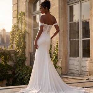 May include: A white off-the-shoulder wedding dress with a fitted bodice and a flowing skirt. The dress features lace detailing on the bodice and sleeves, and a long train with lace trim. The bride is standing on a stone patio.
