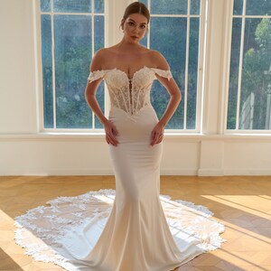 May include: A white off-the-shoulder wedding gown with a fitted bodice and a mermaid skirt. The bodice features intricate lace detailing, and the skirt flows into a long train with lace trim. The dress is displayed in a bright room with large arched windows.