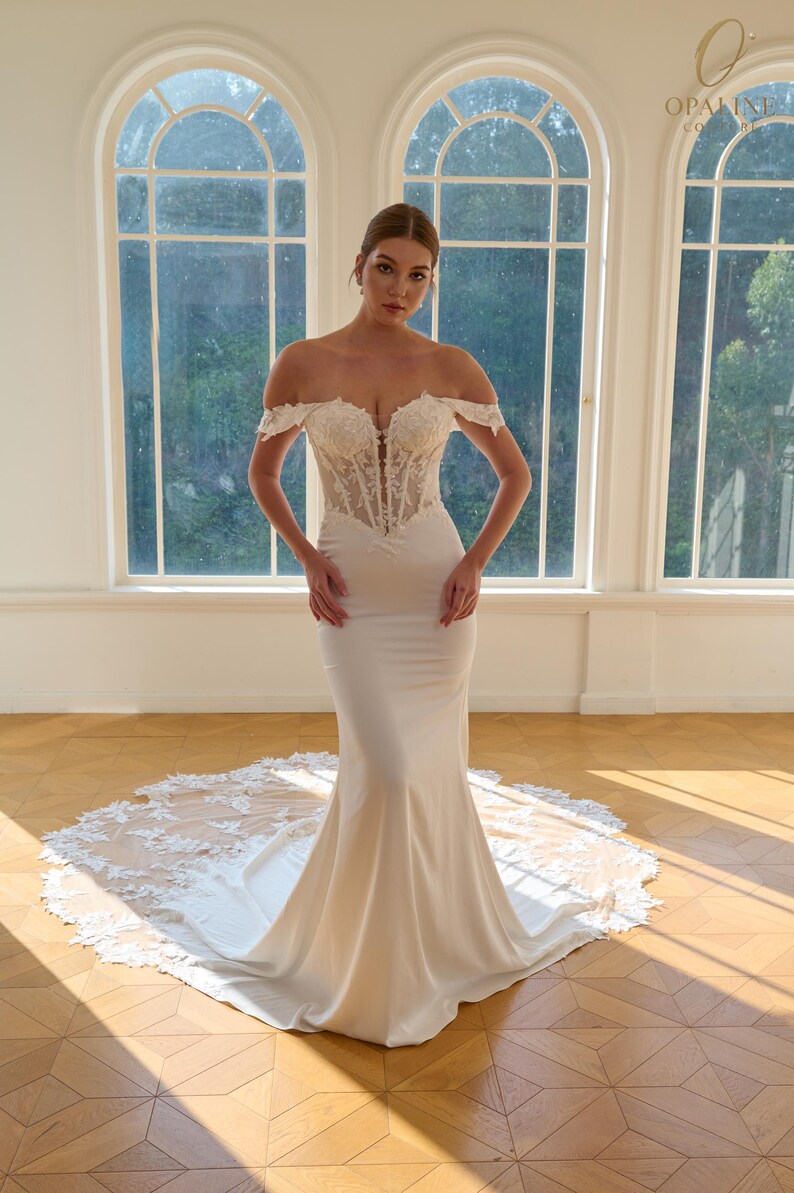 May include: A white off-the-shoulder wedding gown with a fitted bodice and a mermaid skirt. The bodice features intricate lace detailing, and the skirt flows into a long train with lace trim. The dress is displayed in a bright room with large arched windows.