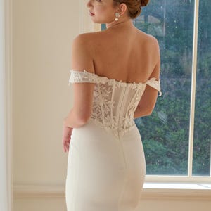 May include: A white off-the-shoulder wedding dress with a fitted bodice and a long, flowing skirt. The bodice features intricate floral lace detailing. The dress is elegant and simple, perfect for a bride.