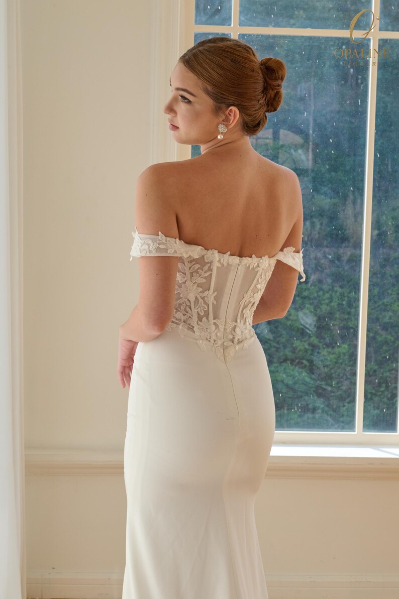May include: A white off-the-shoulder wedding dress with a fitted bodice and a long, flowing skirt. The bodice features intricate floral lace detailing. The dress is elegant and simple, perfect for a bride.