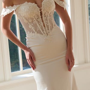 May include: Ivory off-the-shoulder wedding gown with a fitted silhouette. The bodice features intricate floral lace detailing and a sheer panel. The dress has a flowing skirt and is set against a bright window background. The Opaline Couture logo is visible.