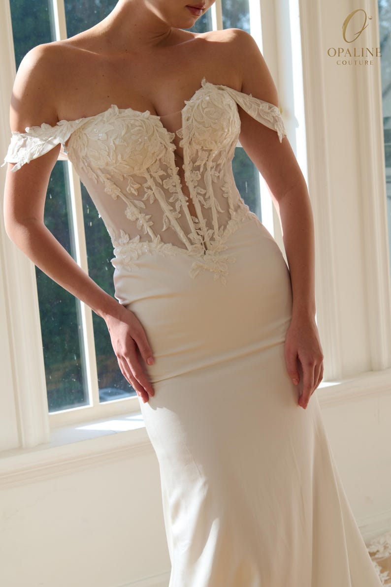 May include: Ivory off-the-shoulder wedding gown with a fitted silhouette. The bodice features intricate floral lace detailing and a sheer panel. The dress has a flowing skirt and is set against a bright window background. The Opaline Couture logo is visible.