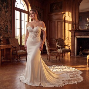 May include: A white off-the-shoulder wedding gown with a fitted bodice and a flowing skirt that extends into a train with lace detailing. The dress is made of a satin-like material. The background is a room with wooden paneling and antique furniture.