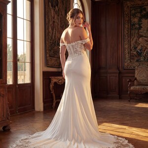 May include: A white off-the-shoulder wedding gown with a fitted bodice and a flowing mermaid skirt. The dress features lace detailing on the bodice and a long train with lace trim. The image is set in a room with wooden furniture and tapestries.