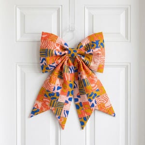 May include: A large decorative bow made of quilted fabric featuring a patchwork pattern of squares in shades of orange, pink, mustard yellow, and blue. The fabric displays various abstract shapes, wavy lines, and geometric motifs throughout the design.