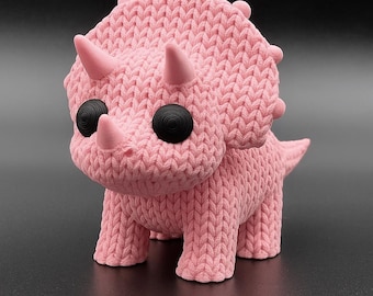May include: A pink triceratops figurine with a knitted texture. The dinosaur has three horns, black circular eyes, and a small tail. The figurine is standing on a dark surface.