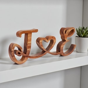 Puede incluir: Letrero decorativo de madera que deletrea "J & E" en una fuente cursiva. El "&" está representado por una forma de corazón. El letrero está hecho de madera de color marrón claro y se muestra en un estante blanco. A la derecha hay una pequeña suculenta en maceta.