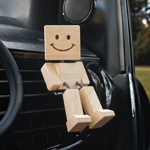 May include: A wooden figure with a smiling face sits on a black car dashboard. The figure has a square head, rectangular body, and jointed limbs. The background shows a speaker and a glimpse of a green, grassy area.