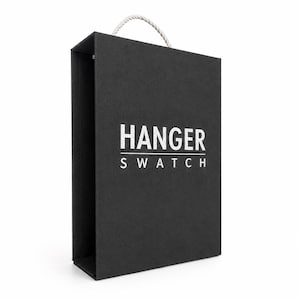 Puede incluir: Una caja rectangular negra con un asa de cuerda blanca en la parte superior. El frente de la caja presenta el texto blanco HANGER SWATCH impreso en una fuente sans-serif en negrita. La caja tiene un acabado mate y un diseño estructurado.