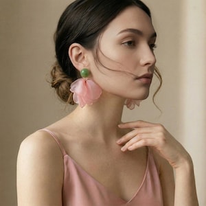 Puede incluir: Pendientes de flores rosas con un perno verde. Los pendientes están hechos de una tela delicada y transparente, creando un aspecto suave y femenino. Estos pendientes son una pieza llamativa.