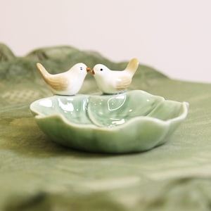 May include: A ceramic dish with a leaf shape, holding two small white birds with tan wings. The birds are positioned facing each other. The dish is a light green color.
