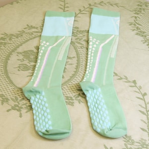May include: A pair of light green socks with a light blue top section. The socks have a dotted pattern on the bottom and white and lavender stripes down the sides. The socks are designed for comfort and support.