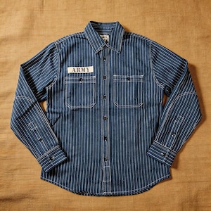 Puede incluir: Una camisa azul a rayas con la palabra "ARMY" en un parche sobre el bolsillo izquierdo del pecho. La camisa tiene dos bolsillos en el pecho, un cuello y mangas largas con puños abotonados. Rayas verticales blancas recorren la camisa.