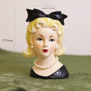Peut inclure: Un vase tête en céramique de style vintage, représentant une femme blonde aux lèvres rouges et un nœud noir. Le vase mesure 13,5 cm de haut, 8 cm de large et 6,5 cm de profondeur. Il comprend un collier de perles et des boucles d'oreilles. Le nœud mesure 3,4-4,3 cm.