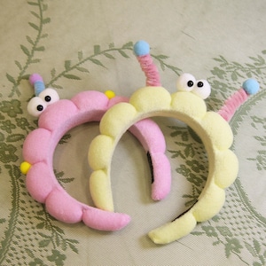 May include: Two whimsical headbands, one pink and one yellow, each shaped like a caterpillar. They feature large googly eyes, pink and blue antennae, and yellow accents. The headbands are designed for fun and costume wear.